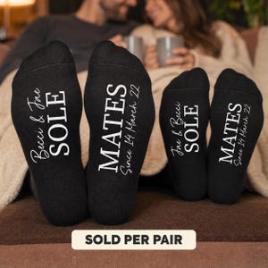 May include: Two pairs of black socks with white text printed on the soles. The first pair reads Becci & Jae SOLE and MATES Since 14 March 22. The second pair reads Jae & Becci SOLE and MATES Since 14 March 22 in a matching font style.