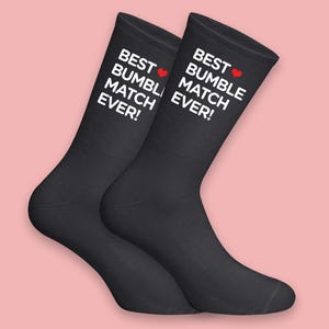 Puede incluir: Dos calcetines negros con el texto blanco "BEST BUMBLE MATCH EVER!" y un corazón rojo. Los calcetines se muestran sobre un fondo rosa claro.