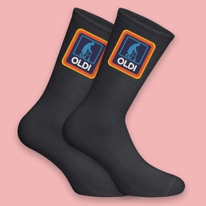 Puede incluir: Dos calcetines negros con la palabra "OLDI" y un logotipo de una persona con andador. El logotipo está en un cuadrado azul con un borde naranja. Fondo rosa.