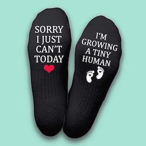 Könnte beinhalten: Schwarze Socken mit weißem Text: "SORRY I JUST CAN'T TODAY" mit rotem Herz und "I'M GROWING A TINY HUMAN" mit weißen Fußabdrücken. Diese Socken sind ein lustiges Geschenk für werdende Eltern.