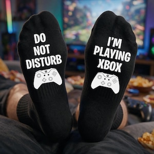 May include: A pair of black socks featuring white text and graphics. The left sock displays the text DO NOT DISTURB above a white game controller icon. The right sock displays the text I'M PLAYING XBOX above a white game controller icon.