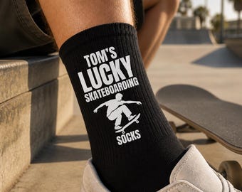 Op de afbeelding: Een zwarte sok met witte tekst die TOM'S LUCKY SKATEBOARDING SOCKS leest en een witte afbeelding van een persoon op een skateboard. De sok heeft een geribbelde textuur en wordt gedragen aan het onderbeen boven een lichtgekleurde sneaker.