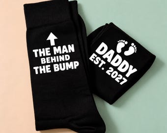 Op de afbeelding: Een paar zwarte sokken met witte tekst. Eén sok heeft een pijl die naar boven wijst en de tekst "THE MAN BEHIND THE BUMP". De andere sok toont twee babyvoetjes en de tekst "DADDY EST. 2027" in een vetgedrukt, schreefloos lettertype.