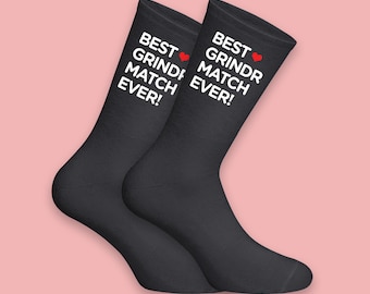 May include: Two black socks with the text "BEST GRINDR MATCH EVER!" in white, and a red heart symbol. The socks are shown against a light pink background.