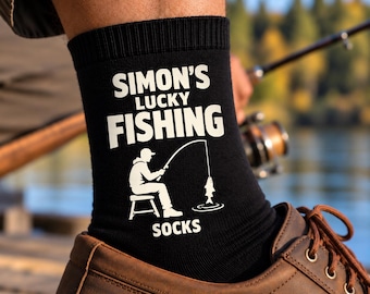 May include: A black crew sock featuring white text that reads SIMON'S LUCKY FISHING SOCKS. Below the text is a white graphic illustration of a person sitting on a stool while fishing with a rod and line, with a fish caught on the end of the line.