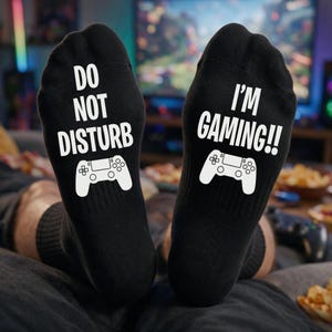 Peut inclure: Une paire de chaussettes noires avec du texte blanc et un graphique de manette de jeu blanc. La chaussette gauche porte l'inscription DO NOT DISTURB et la droite I'M GAMING!!, toutes deux au-dessus d'une illustration de manette de jeu.