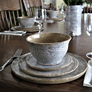 May include: A rustic ceramic bowl and plates set in neutral tones, arranged on a dark wooden table. The set includes a large bowl, a medium plate, and a smaller plate. A fork and wine glasses are also visible, along with a vase of purple flowers.