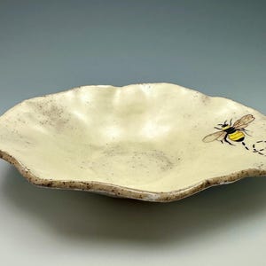 May include: A handmade, off-white ceramic serving dish with a unique, organic shape. The dish features a hand-painted bee with yellow and black details, and a dotted flight path. The rim has a speckled, brown edge.