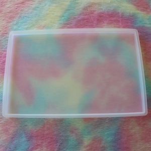 May include: A rectangular, translucent silicone mold with a raised border. The mold is empty, and the interior is a soft, pastel color. The background is a colorful, tie-dye pattern in shades of pink, blue, and yellow.