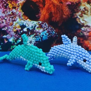 May include: Two beaded dolphin figurines. One is turquoise, the other is light blue. Both have black eyes and white bellies. The dolphins are on a blue surface, with a colorful coral reef background. Each dolphin has a clear, beaded loop.