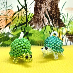 May include: Two beaded turtle figurines. One is a solid green, the other is a translucent green. Both have black eyes and clear legs. The turtles are on a yellow surface with a painted background of trees and foliage.