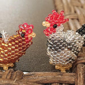 May include: Two beaded chicken figurines. One is gold and brown, the other is silver, black, and red. The chickens have detailed beadwork, with red combs and black eyes. They are placed on a dark surface.