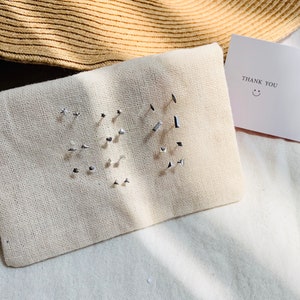 Pu&ograve; includere: Un set di orecchini d'argento esposti su una pochette in tessuto bianco. Gli orecchini sono disposti in una variet&agrave; di forme, tra cui stelle, quadrati e cerchi. Un piccolo biglietto bianco con il testo "THANK YOU" e un'emoticon sorridente &egrave; visibile sullo sfondo.