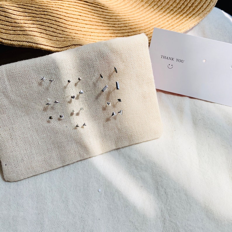 Pu&ograve; includere: Un set di orecchini d'argento esposti su una pochette in tessuto bianco. Gli orecchini sono disposti in una variet&agrave; di forme, tra cui stelle, quadrati e cerchi. Un piccolo biglietto bianco con il testo "THANK YOU" e un'emoticon sorridente &egrave; visibile sullo sfondo.