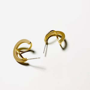 May include: A pair of gold-toned hoop earrings with a unique twisted design. The earrings are made of metal and have a simple, modern style.