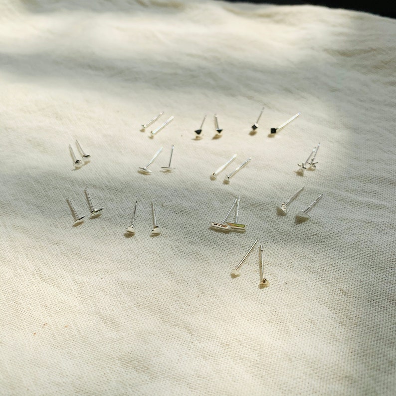 Pu&ograve; includere: Una collezione di orecchini d'argento con varie forme geometriche, tra cui quadrati, triangoli e cerchi, disposti su uno sfondo di tessuto bianco.
