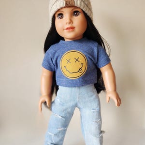 May include: A doll wearing a beige knit beanie with a faux fur pom-pom, a blue t-shirt with a yellow smiley face graphic, and light blue ripped jeans. The doll is also wearing black and white sneakers.