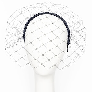 May include: A black netting veil attached to a black headband. The netting has a diamond pattern and is draped over a white mannequin head. The headband is made of a smooth, shiny material.