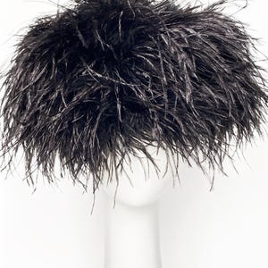May include: A black feather hat, likely an accessory for special occasions, is displayed on a white mannequin head. The hat is densely packed with dark feathers, creating a voluminous, textured appearance. The feathers have a slight sheen.