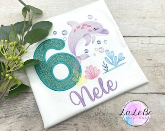 May include: White t-shirt with an embroidered design featuring a purple dolphin, bubbles, and aquatic plants. A teal number 6 with glitter detail is on the left. The name "Nele" is written in purple script.