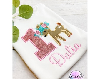 May include: White long-sleeve shirt featuring a glittery pink number 1 and the name "Dalia" embroidered in pink. A brown deer with a floral crown is embroidered on the shirt. The shirt is from LaLeBe Creations.