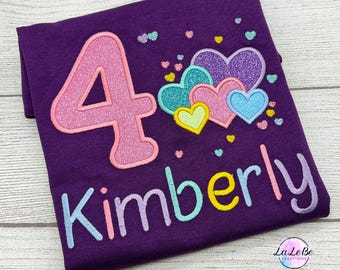 Könnte beinhalten: Ein dunkelviolettes T-Shirt mit einer großen, glitzernden rosa Zahl Vier, einer Ansammlung von bunten Glitzerherzen in Violett, Türkis, Rosa, Blau und Gelb sowie dem Namen Kimberly, der in hellblauen, rosa, violetten und gelben Buchstaben gestickt ist.