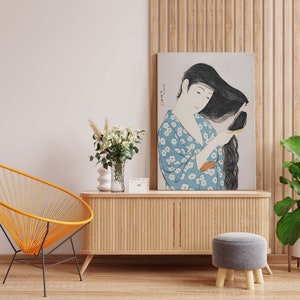 Könnte beinhalten: Ein Holzschränkchen mit einer hellen Holzoptik und vertikalen Lamellen. Über dem Schränkchen hängt ein gerahmtes Bild einer Frau mit langen schwarzen Haaren in einem blau-weiß gemusterten Kimono. Im Vordergrund stehen ein gelber Stuhl und ein grauer Hocker.