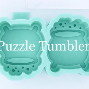 May include: Two green silicone molds for making resin crafts. The molds are shaped like a cauldron and a round object with a rim. The molds are on a white surface next to a ruler. The text "Puzzle Tumblers" is printed on the molds.