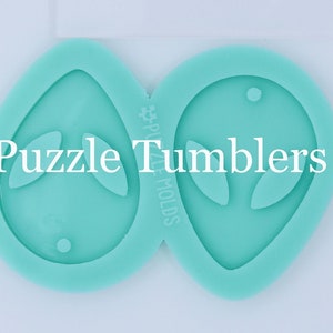 May include: Two teal silicone molds shaped like alien heads with a hole at the top for hanging. The molds are next to a ruler with inch and centimeter markings. The text "Puzzle Tumblers" is printed on the molds.