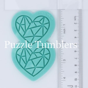 May include: A teal silicone mold with two heart shapes. Each heart has a geometric design of intersecting lines creating a diamond pattern. The mold is 2 inches tall and is next to a ruler for size reference.