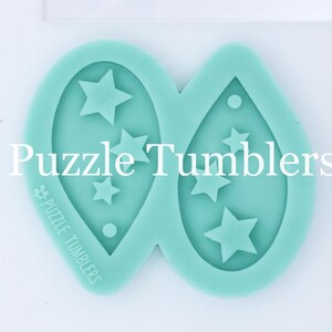 K&ouml;nnte beinhalten: Zwei t&uuml;rkisfarbene Silikonformen zum Herstellen von Ohrringen. Jede Form hat eine Tropfenform mit einem Loch oben und einem Design aus f&uuml;nf Sternen im Inneren. Die Formen sind mit "Puzzle Tumblers" beschriftet.