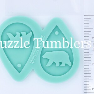 May include: Two teal blue silicone molds for making resin earrings. One mold features a bear silhouette and the other features two bears holding a heart. The molds are on a white surface next to a ruler.