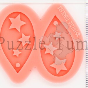 K&ouml;nnte beinhalten: Eine rosa Silikonform zum Herstellen von Ohrringen. Die Form hat zwei tropfenf&ouml;rmige Vertiefungen mit einem Sternmuster. Die Form ist 5 cm lang und 2,5 cm breit.