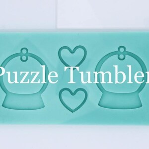 May include: A light blue silicone mold with two round shapes and two heart shapes. The mold is labeled "Puzzle Tumblers".