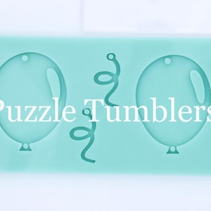 May include: A light green silicone mold with two balloon shapes and a curly ribbon shape. The mold is 6 inches long and is next to a ruler for size reference. The text "Puzzle Tumblers" is printed on the mold.