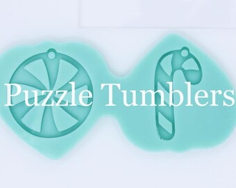 May include: Two silicone molds for making candy ornaments. One mold is shaped like a candy cane and the other is shaped like a peppermint swirl. The molds are light blue and are sitting on a white surface. The text "Puzzle Tumblers" is printed on the surface.