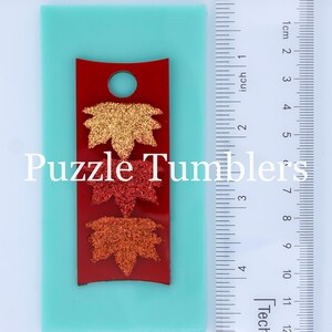 May include: A red silicone mold with three leaf shapes. The leaves are in shades of orange, red, and gold with glitter. The mold is on a turquoise surface with a ruler for scale.