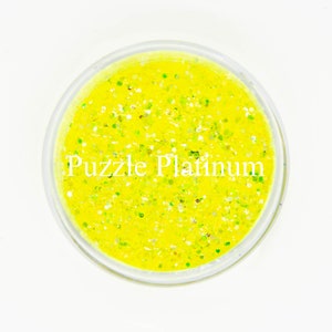 May include: A clear, round container filled with bright yellow glitter. The glitter has a mix of sizes and colors, including green and silver. The text "Puzzle Platinum" is printed in white across the container.