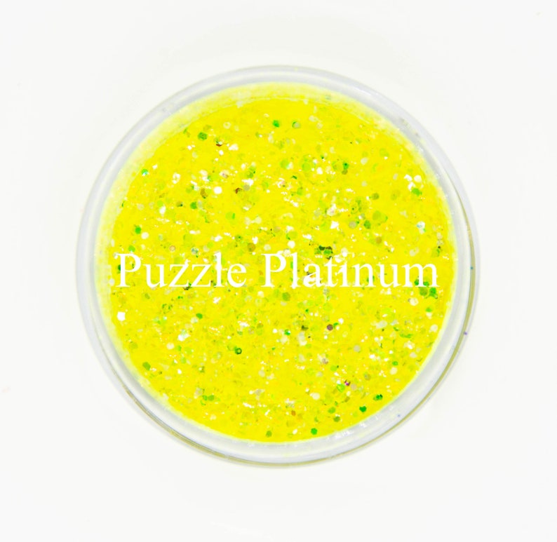 May include: A clear, round container filled with bright yellow glitter. The glitter has a mix of sizes and colors, including green and silver. The text "Puzzle Platinum" is printed in white across the container.