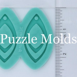 May include: Two teal blue silicone molds for making earrings. Each mold is shaped like a teardrop with a smaller teardrop shape inside. The molds are 2 inches long and are next to a ruler for size reference. The text "#PuzzleMakers" is printed on the side of the molds.