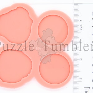 May include: A peach-colored silicone mold with four cavities: two circular and two irregularly shaped. The mold has the text "Puzzle Tumblers" printed on it. A ruler with inch and centimeter measurements is visible.