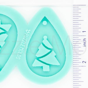 May include: A teal silicone mold with two teardrop-shaped cavities. Each cavity features a Christmas tree design with a wavy line pattern. The mold is labeled "#PUZZLE MOLDS".