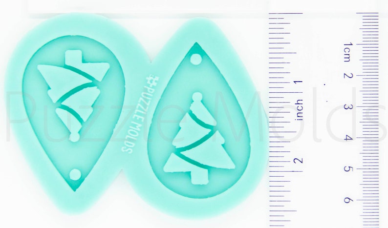 May include: A teal silicone mold with two teardrop-shaped cavities. Each cavity features a Christmas tree design with a wavy line pattern. The mold is labeled "#PUZZLE MOLDS".