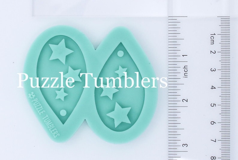 K&ouml;nnte beinhalten: Zwei t&uuml;rkisfarbene Silikonformen zum Herstellen von Ohrringen. Jede Form hat eine Tropfenform mit einem Loch oben und einem Design aus f&uuml;nf Sternen im Inneren. Die Formen sind mit "Puzzle Tumblers" beschriftet.