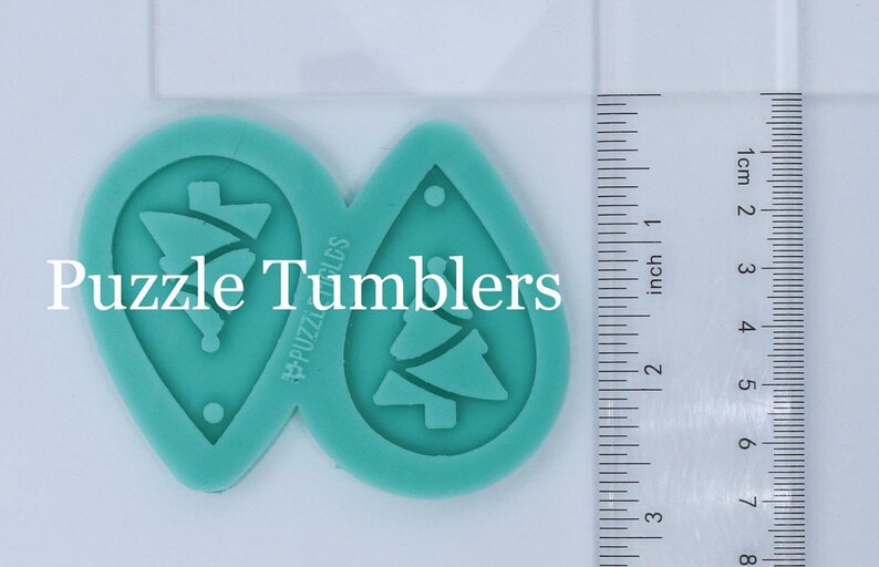 May include: Two teal-colored silicone molds for making earrings. Each mold is shaped like a teardrop and features a Christmas tree design. The molds are approximately 2 inches in length.