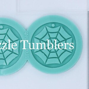 May include: Two teal blue silicone molds for making resin crafts. Each mold is shaped like a circle and features a raised design of a spider web. The molds are labeled "Puzzle Tumblers" and "PU MOLDS".