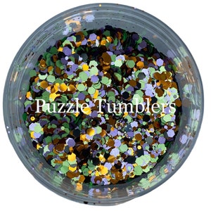 May include: A close-up of a jar filled with green, purple, gold, and black glitter. The glitter is in various shapes and sizes. The jar is labeled "Puzzle Tumblers".