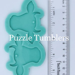 May include: Two teal blue silicone moulds shaped like cow heads. The moulds are approximately 7.5 cm tall and 6.5 cm wide. The moulds are on a white surface next to a ruler.