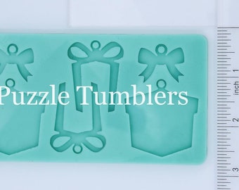 May include: A teal blue silicone mold with four cutouts for making resin ornaments. The mold features a gift box, a bow, and two different bow designs. The text "Puzzle Tumblers" is printed on the mold.