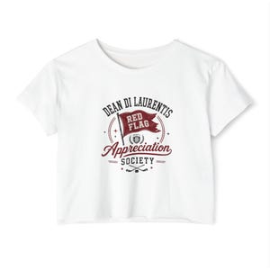 May include: White crop top with a maroon and white graphic. The graphic reads "DEAN DI LAURENTIS RED FLAG Appreciation SOCIETY" with a flag and hockey sticks. The shirt is made of a soft material.